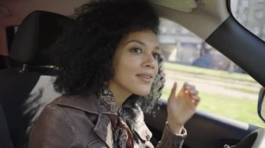 Araba süren genç bir Afro-Amerikan kadının portresi. Kahverengi deri ceketli kıvırcık saçlı bir esmer arabanın içinde direksiyon başında poz veriyor. Kapatın. Yavaş çekim.