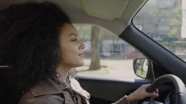 Araba süren genç bir Afro-Amerikan kadının portresi. Kahverengi deri ceketli kıvırcık saçlı bir esmer arabanın içinde direksiyon başında poz veriyor. Kapatın. Yavaş çekim.