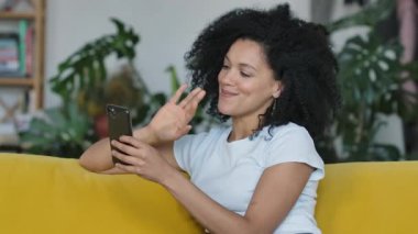 Genç bir Afro-Amerikan kadının portresi akıllı telefonuyla video görüşmesi yapıyor. Sarı koltukta oturan kıvırcık saçlı esmer, parlak bir ev odasında oturuyor. Kapatın. Yavaş çekim.