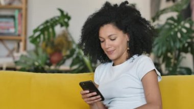 Genç bir Afro-Amerikalı kadının portresi. Telefonuna mesaj atıyor. Sarı koltukta oturan kıvırcık saçlı esmer, parlak bir ev odasında oturuyor. Kapatın. Yavaş çekim.