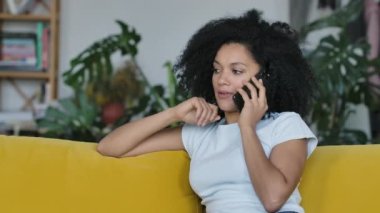 Cep telefonu için konuşan genç bir Afrikalı Amerikalı kadının portresi. Sarı koltukta oturan kıvırcık saçlı esmer, parlak bir ev odasında oturuyor. Kapatın. Yavaş çekim.