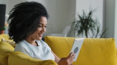 Genç bir Afro-Amerikan kadının portresi dijital tabletindeki bir video görüşmesinde konuşuyor. Sarı koltukta oturan kıvırcık saçlı esmer, parlak bir ev odasında oturuyor. Kapatın. Yavaş çekim.