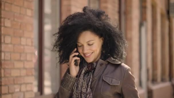 年轻的非洲裔美国女人用智能手机说话的肖像。穿着皮夹克卷曲头发的布鲁内特在模糊的砖楼背景下沿街走着。慢动作靠近点. — 图库视频影像