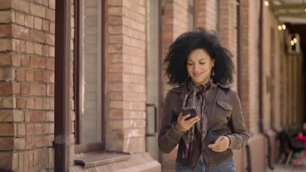 年轻的非洲裔美国女人用智能手机说话的肖像。穿着皮夹克卷曲头发的布鲁内特在漂亮的砖楼背景下沿街走着。慢动作. — 图库视频影像