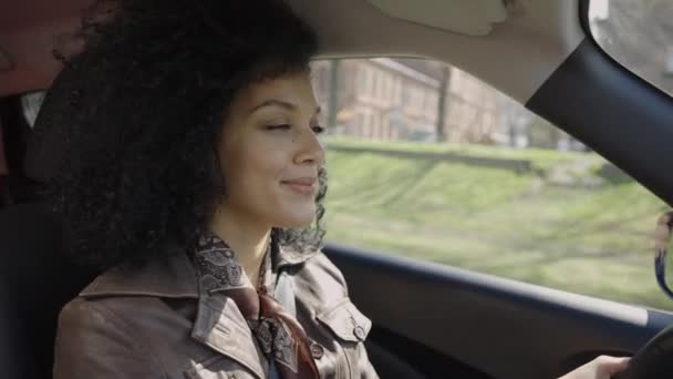 Retrato de una joven y elegante mujer afroamericana conduciendo un coche. Una morena con el pelo rizado en una chaqueta de cuero marrón posa detrás del volante dentro de un coche. De cerca. Movimiento lento. — Vídeo de stock