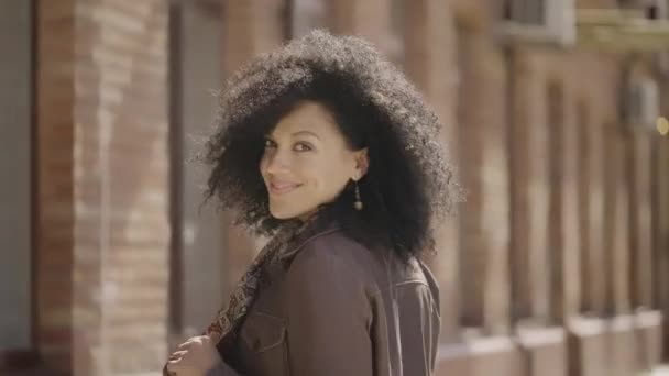 Achteraanzicht van de jonge Afro-Amerikaanse vrouw die door de straat loopt, zich omdraait en glimlacht. Brunette in bruin leren jasje poseert tegen de achtergrond van wazig bakstenen gebouw. Langzame beweging. Sluiten.. — Stockvideo
