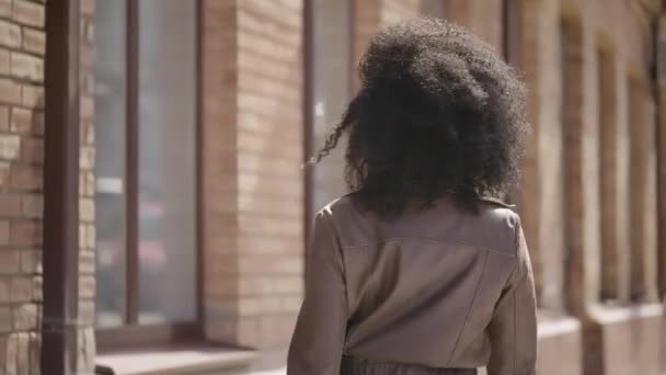 Rear view of young African American woman walking down street, turning around and smiling. Brunette in brown leather jacket poses against background of blurred brick building. Slow motion. Close up. — 图库视频影像