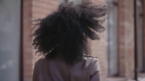 Rear view of young African American woman walking down street, turning around and smiling. Brunette in brown leather jacket poses against background of blurred brick building. Slow motion. Close up. — 图库视频影像