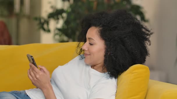 젊은 아프리카 계 미국인 여성 이 핸드폰으로 문자를 하는 모습. 밝은 집 방에 노란색 소파에 곱슬곱슬 한 머리의 브루넷. 느린 동작. 닫아. — 비디오