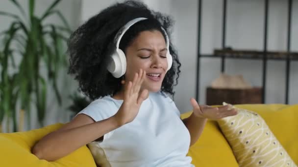 Porträtt av en ung afroamerikansk kvinna njuter av musik med stora vita hörlurar. Brunett med lockigt hår sittande på gul soffa i ett ljust hemrum. Närbild. Långsamma rörelser. — Stockvideo