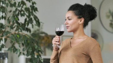 Genç Afrikalı Amerikalı kadının cam kadehten kırmızı şarap içişi. Şirin melez kadın ışık odasının arka planında içkinin tadını çıkarıyor. Kapatın. Yavaş çekim.