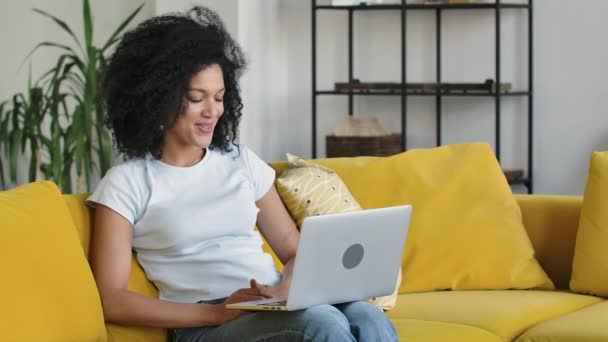 Porträt einer jungen Afroamerikanerin bei einem Videoanruf auf einem tragbaren Laptop. Brünette mit lockigem Haar sitzt auf einem gelben Sofa in einem hellen Wohnzimmer. Aus nächster Nähe. Zeitlupe. — Stockvideo