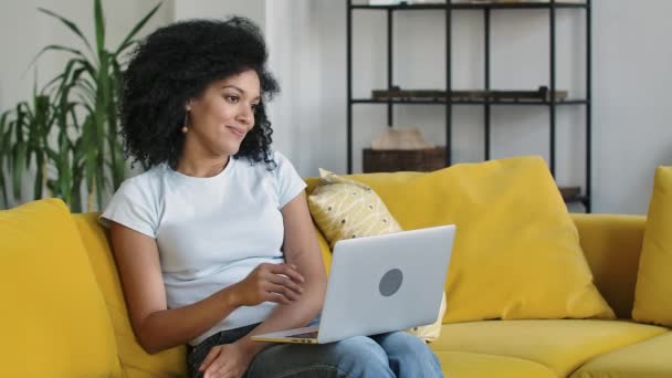 Portret młodej Afroamerykanki rozmawiającej przez telefon na przenośnym laptopie. Brunetka z kręconymi włosami siedząca na żółtej kanapie w jasnym salonie. Zamknij drzwi. Zwolniony ruch. — Wideo stockowe