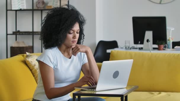 Ritratto di una giovane donna afroamericana che digita sulla tastiera di un computer portatile. Bruna con capelli ricci seduta su un divano giallo in una luminosa stanza di casa. Chiudete. Rallentatore. — Video Stock