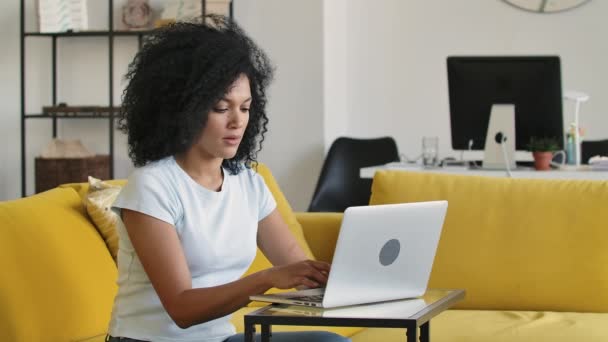 Porträt einer jungen Afroamerikanerin beim Tippen auf der Laptop-Tastatur. Brünette mit lockigem Haar sitzt auf einem gelben Sofa in einem hellen Wohnzimmer. Aus nächster Nähe. Zeitlupe. — Stockvideo