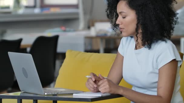 Porträt einer jungen Afroamerikanerin spricht auf Videokonferenz mit Laptop und macht sich Notizen in Notizbuch. Brünette mit lockigem Haar sitzt auf einem gelben Sofa im Wohnzimmer. Aus nächster Nähe. Zeitlupe. — Stockvideo