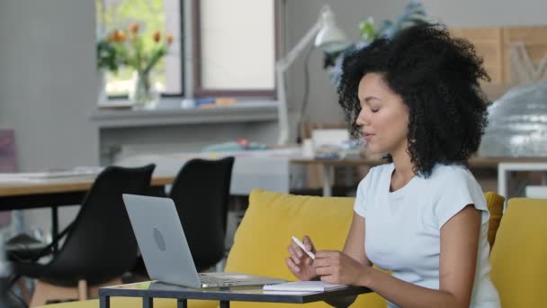 Porträt einer jungen Afroamerikanerin spricht auf Videokonferenz mit Laptop und macht sich Notizen in Notizbuch. Brünette mit lockigem Haar sitzt auf einem gelben Sofa im Wohnzimmer. Aus nächster Nähe. Zeitlupe. — Stockvideo