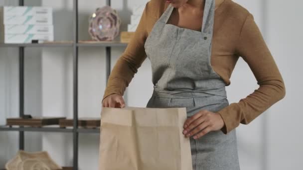 Chica joven recoge una bolsa de papel con su pedido en línea en una tienda. Una linda mujer afroamericana está empacando productos para un cliente en el fondo de la sala de luz. De cerca. Movimiento lento. — Vídeo de stock