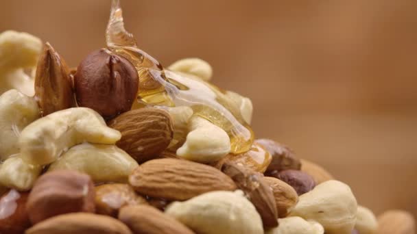 Ein Rinnsal goldenen Honigs fließt in Zeitlupe einen Haufen Nüsse hinunter. Honig tropft und umhüllt die Mischung aus Mandeln, Cashewnüssen und Haselnüssen. Detailaufnahme von Nüssen in süßem Sirup. — Stockvideo