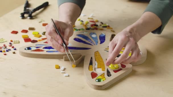 Las manos masculinas crean mosaicos con pinzas y pequeñas piezas brillantes de vidrio y cerámica. Artesano maestro haciendo patrón de mariposa de colores en un taller creativo. De cerca. Movimiento lento. — Vídeos de Stock