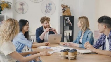 Bir grup genç erkek ve kadın modern ofiste masada oturuyorlar ve müzakere toplantısında iş meselelerini tartışıyorlar. Nitelikli çalışanlar başlangıç pazarlama stratejisi geliştiriyor. Yavaş çekim.