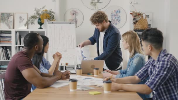 Jonge mannelijke teamleider staat vlak bij het bestuur en presenteert een nieuw opstartproject. Gemengde race collega 's bespreken werkkwesties tijdens de onderhandelingsvergadering. Langzame beweging. — Stockvideo