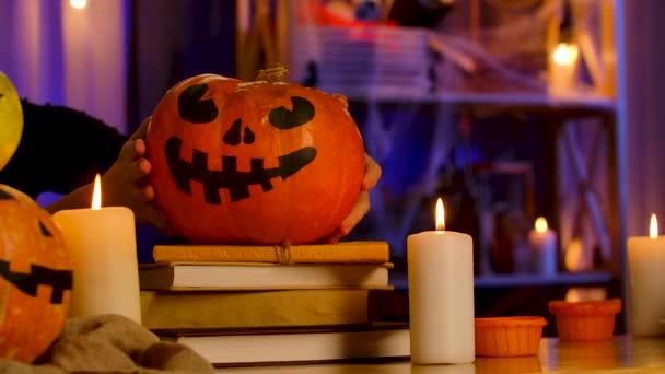 Em um quarto escuro decorado para o Halloween, as velas estão queimando, as mãos das meninas colocam uma abóbora com um rosto assustador pintado em uma pilha de livros e guardá-lo. O fundo está desfocado. Fechar. — Vídeo de Stock