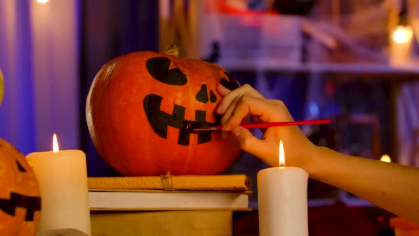 The girls hand with a brush paints a scary face on a pumpkin. The pumpkin lies on the books next to burning candles in a dark, Halloween decorated room. The background is blurred. Halloween. Close up — Stock Video