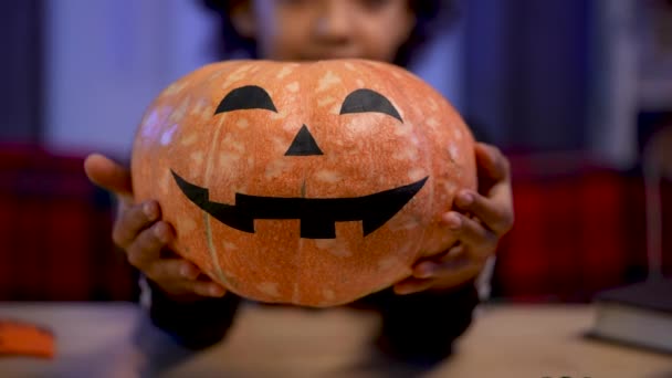 Malá Afroameričanka ve slavnostním kostýmu drží oranžovou dýni s namalovanou usměvavou tváří. Dítě sedí u stolu v pokoji vyzdobeném na Halloweenskou noc. Zavřít. Zpomalený pohyb. — Stock video