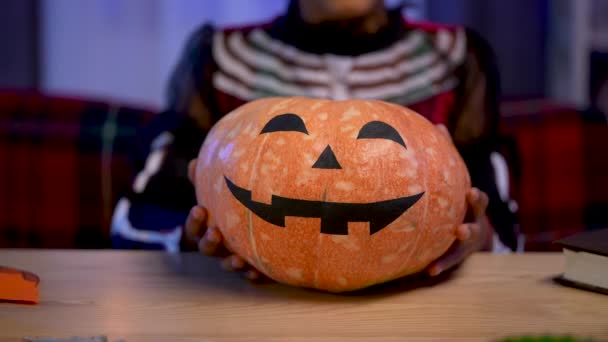 En liten afroamerikansk flicka i festlig dräkt håller en orange pumpa med målat leende ansikte. Barnet sitter vid ett bord i rum inrett för Halloween natten. Närbild. Långsamma rörelser. — Stockvideo