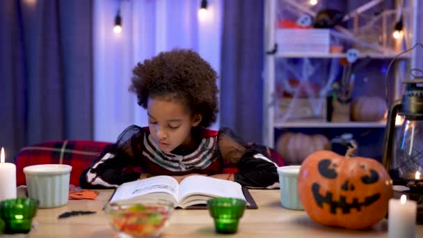 Klein Afrikaans Amerikaans meisje in feestelijk kostuum leest boek met enthousiasme. Kind zit aan tafel in kamer ingericht voor de Halloween avond. Enge en leuke vakantie met het gezin. Sluit maar af. Langzame beweging. — Stockvideo