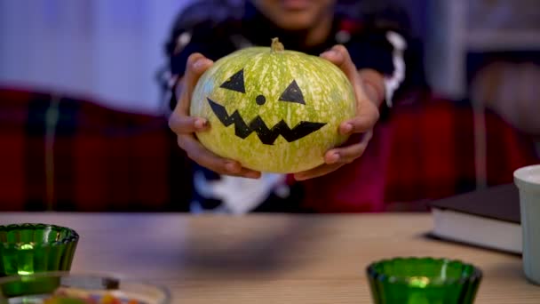 Little African American girl in festive costume is holding an pumpkin with painted smiling face. Child sits at a table in room decorated for the Halloween night. Close up. Slow motion. — Stock Video