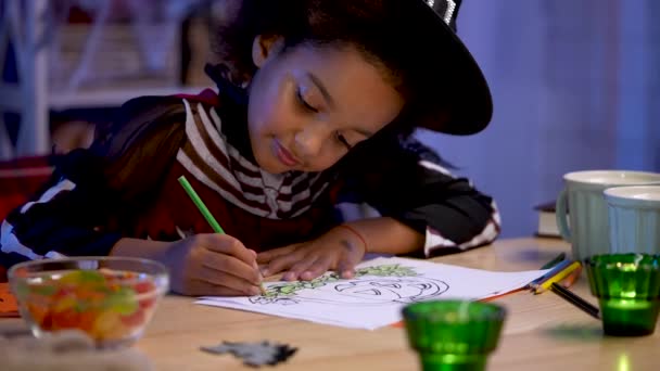 En liten afroamerikansk flicka i festlig dräkt och häxhatt ritar pumpa med pennor. Barnet sitter vid bordet i rummet inrett för Halloween natten. Närbild. Långsamma rörelser. — Stockvideo