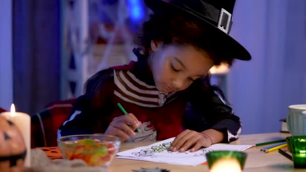 Mała Afroamerykanka w świątecznym kostiumie i kapeluszu czarownicy rysuje dynię ołówkami. Dziecko siedzi przy stole w pokoju urządzonym na noc Halloween. Zamknij drzwi. Zwolniony ruch. — Wideo stockowe