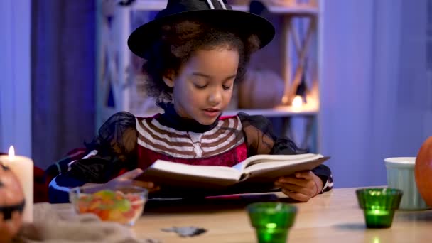 Piccola ragazza afroamericana in costume festivo legge libro con entusiasmo. Il bambino si siede a tavola in camera decorata per la notte di Halloween. Vacanza spaventosa e divertente con la famiglia. Chiudete. Rallentatore. — Video Stock