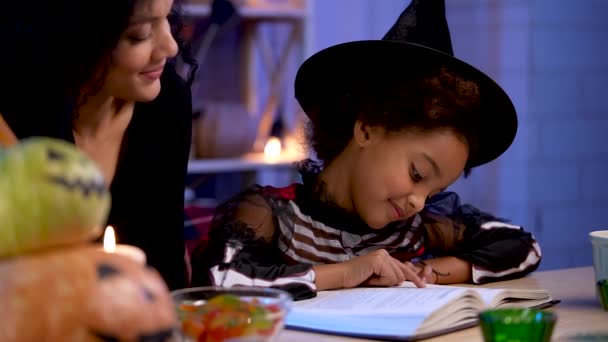 En liten afroamerikansk flicka i festdräkt och häxhatt läser bok. Mamma och dotter sitter vid bordet i ett rum inrett för Halloweenkvällen. Närbild. Långsamma rörelser. — Stockvideo