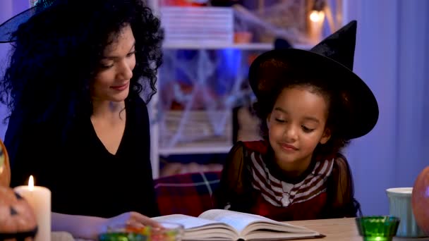お祝いの衣装や魔女の帽子の小さなアフリカ系アメリカ人の女の子は本を読む。母と娘はハロウィンの夜のために装飾された部屋のテーブルに座っています。閉めろ。スローモーション. — ストック動画