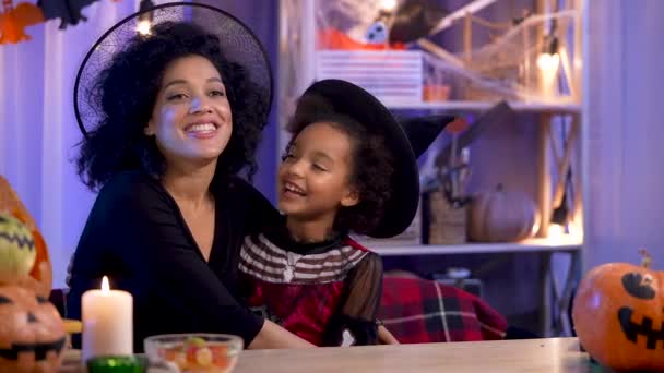 Maman et fille afro-américaine en costume de fête et chapeau de sorcière s'amusent et se câlinent joyeusement. Joyeux famille est assis à table dans la chambre décorée pour la nuit d'Halloween. Ferme là. Mouvement lent. — Video