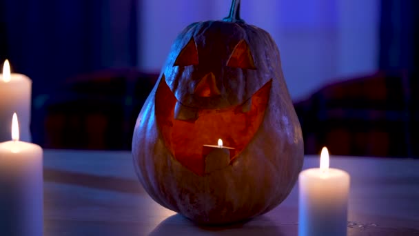 Calabaza tradicional de Halloween con la cara sonriente tallada brilla en la mesa en la oscuridad. Símbolo vegetal naranja de la fiesta en la habitación decorada junto a velas encendidas. De cerca. Movimiento lento. — Vídeos de Stock