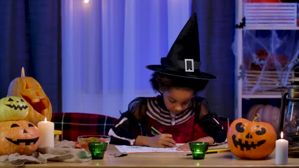 Petite afro-américaine en costume de fête et chapeau de sorcière dessine citrouille avec des crayons. L'enfant est assis à table dans la chambre décorée pour la nuit d'Halloween. Mouvement lent. — Video