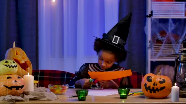 La niñita afroamericana en traje festivo y sombrero de bruja corta la calabaza del papel naranja. El niño se sienta a la mesa en la habitación decorada para la noche de Halloween. Movimiento lento. — Vídeos de Stock