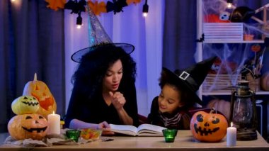 Festivalci kostümlü ve cadı şapkalı küçük Afro-Amerikalı kız kitap okuyordu. Anne ve kızı Cadılar Bayramı için dekore edilmiş bir masada oturuyor. Yavaş çekim.
