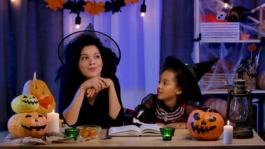 Festival kostümü ve cadı şapkası giymiş anne Afrikalı Amerikalı kızına korkunç bir peri masalı anlatıyor. Mutlu aile Cadılar Bayramı için dekore edilmiş bir masada oturuyor. Yavaş çekim.