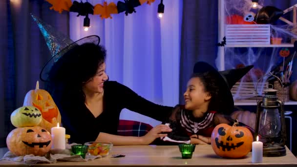 Mama i córka Afroamerykanka w świątecznym kostiumie i kapeluszu czarownicy radośnie się śmieją i bawią. Szczęśliwa rodzina siedzi przy stole w pokoju urządzone na noc Halloween. Zwolniony ruch. — Wideo stockowe