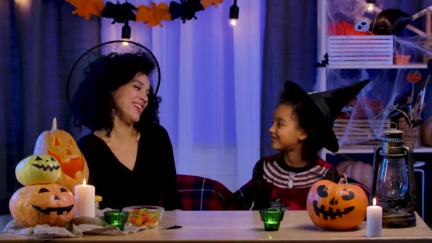 Mamá e hija afroamericanas en traje festivo y sombrero de bruja divirtiéndose y comiendo dulces. La familia feliz se sienta a la mesa en la habitación decorada para la noche de Halloween. Movimiento lento. — Vídeos de Stock