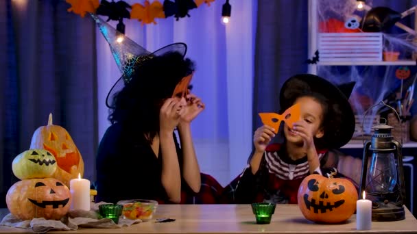 Mamá e hija afroamericanas en traje festivo y sombrero de bruja que se divierten vistiendo máscaras de calabaza naranja. La familia feliz se sienta a la mesa en la habitación decorada para la noche de Halloween. Movimiento lento. — Vídeos de Stock