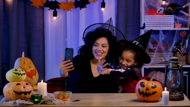 小さな女の子と若い女性魔女の帽子のアフリカ系アメリカ人は、スマートフォンを使用してビデオ通話で通信します。母と娘はハロウィンの夜のために装飾された部屋のテーブルに座っています。スローモーション. — ストック動画