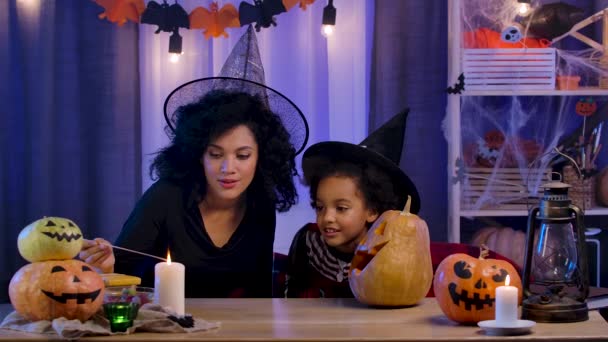 Kleine Mädchen und eine Afroamerikanerin mit Hexenhut entzünden Kerzen in Kürbis mit geschnitztem lächelndem Gesicht. Mutter und Tochter sitzen am Tisch in einem Raum, der für die Halloween-Nacht dekoriert ist. Zeitlupe. — Stockvideo