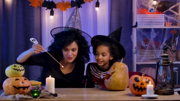 Petite fille et femme afro-américaine en chapeau de sorcière bougie lumineuse en citrouille avec le visage souriant sculpté. Maman et sa fille s'assoient à table dans une chambre décorée pour la nuit d'Halloween. Mouvement lent. — Video