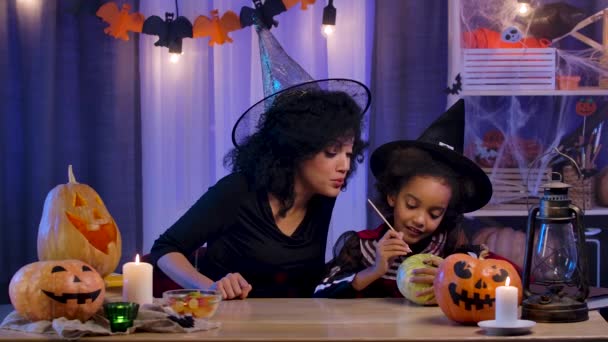 Bambina e donna afroamericana in costume festivo e cappello da strega dipingerà zucca. Mamma e figlia si siedono a tavola in camera decorata per la notte di Halloween. Rallentatore. — Video Stock